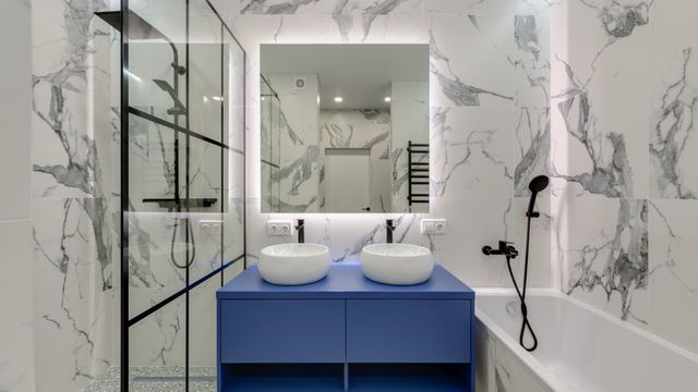 Modern bathroom featuring white marble patterned tiles, a bold blue vanity with two basin sinks, and a bathtub.
