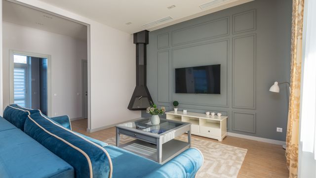 Modern living room interior featuring a blue velvet sofa, a grey coffee table, a low TV console, and a wall-mounted television against a panelled grey wall.