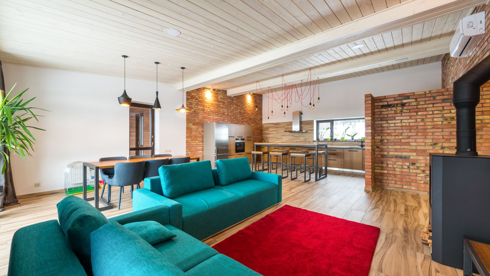 Modern open-plan interior with a teal sofa, red rug, dining table, and an exposed brick kitchen area.