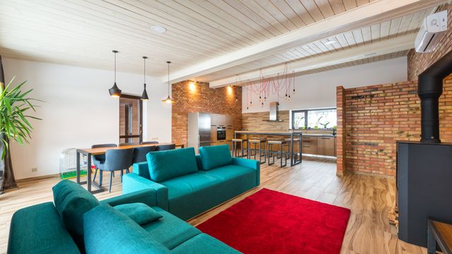Modern open-plan interior with a teal sofa, red rug, dining table, and an exposed brick kitchen area.