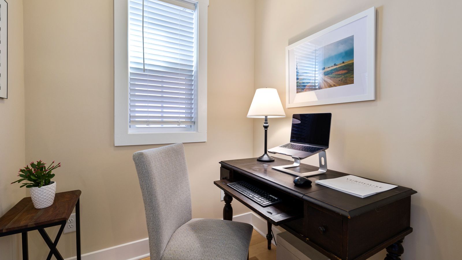 A compact home office workstation featuring a small wooden desk with a laptop on a stand, keyboard tray, mouse, desk lamp and upholstered chair in a small nook.