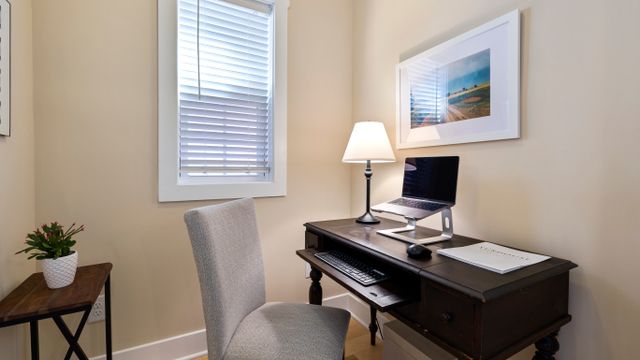 A compact home office workstation featuring a small wooden desk with a laptop on a stand, keyboard tray, mouse, desk lamp and upholstered chair in a small nook.