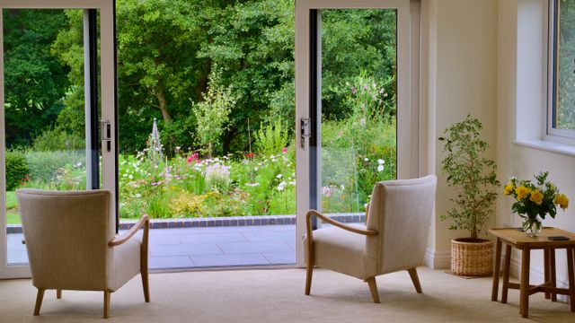 Two armchairs in a bright room facing open glass patio doors that overlook a lush flower garden and trees.