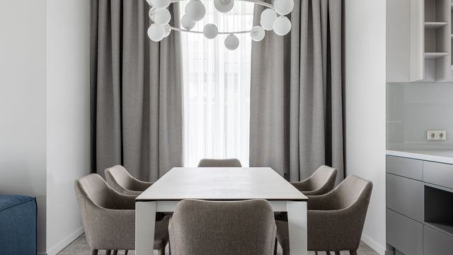 A dining table with grey upholstered chairs positioned before a large curtained window with a modern pendant light.