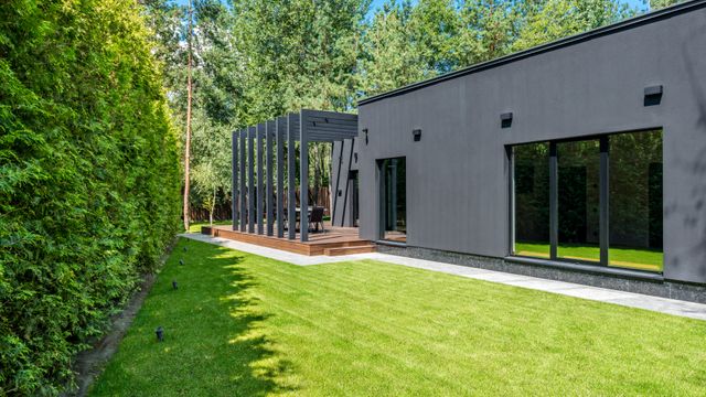 Exterior of a contemporary single-storey charcoal grey house with large windows, timber decking, and a pergola facing a manicured lawn edged by a tall cedar hedge.