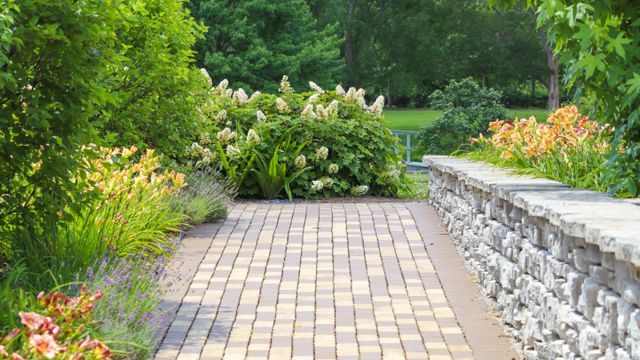 A paved brick path through a garden bordered by flowering plants and a low stone wall.