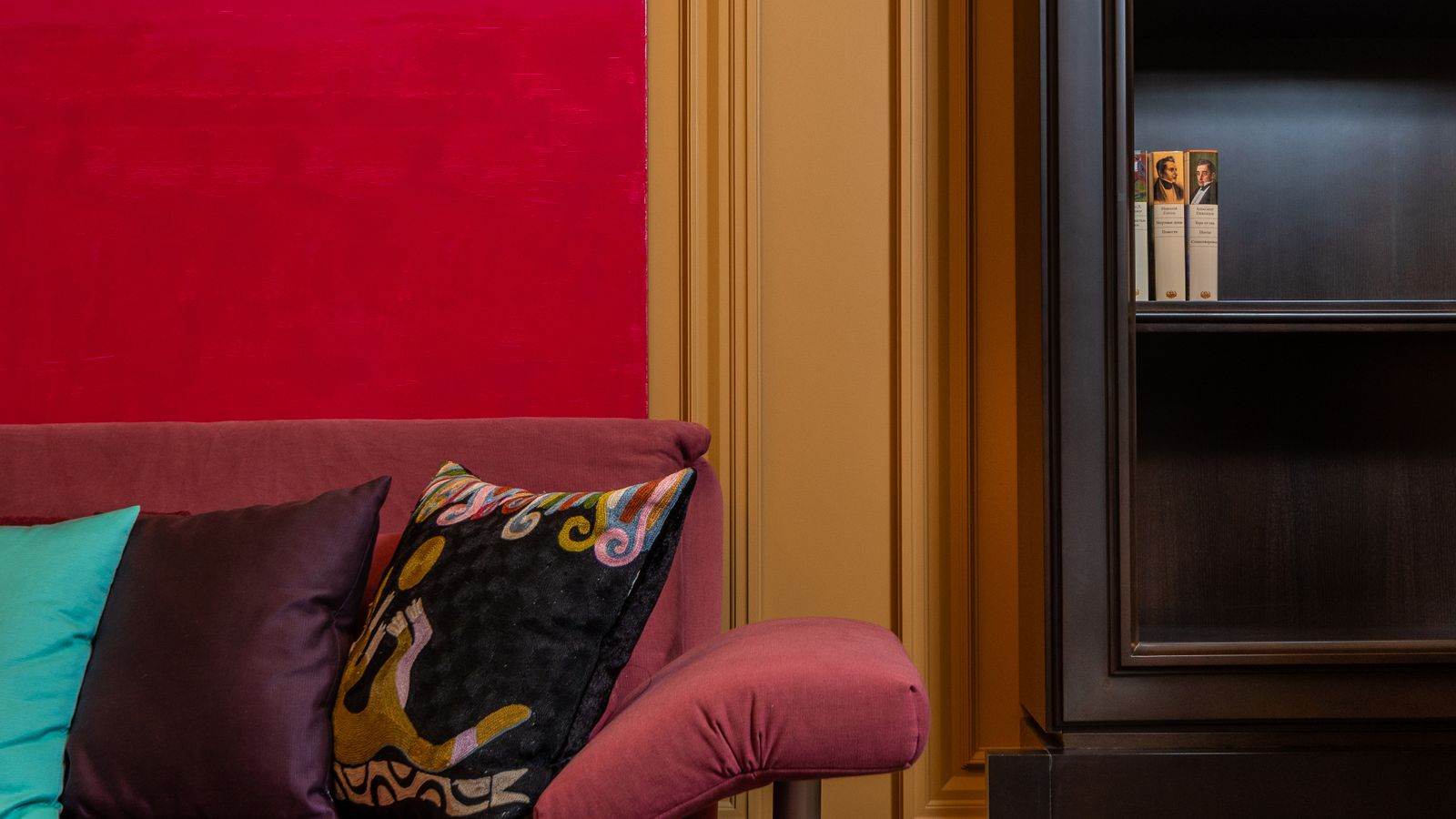 A close-up of a pink sofa with colourful cushions against a bold red feature wall and muted ochre wall moulding beside a dark bookcase.